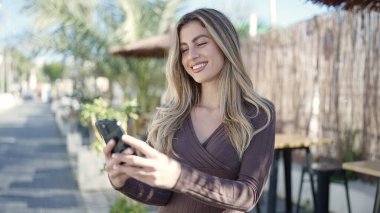 Young blonde woman smiling confident using smartphone at coffee shop terrace