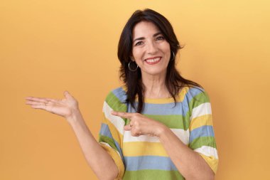 Middle age hispanic woman standing over yellow background amazed and smiling to the camera while presenting with hand and pointing with finger. 