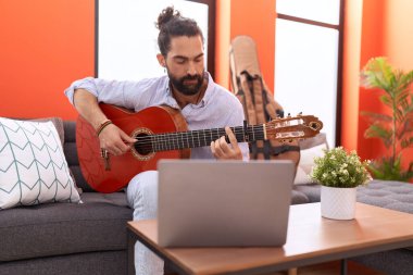 Young hispanic man having online guitar class sitting on sofa at home