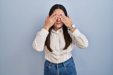 Mavi arka planda dikilen genç Latin kadın yorgunluk ve baş ağrısı için gözlerini ovuşturuyor, uykulu ve yorgun ifade. görme sorunu 