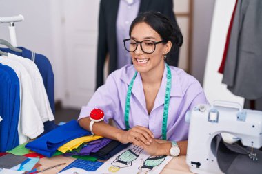 Young beautiful hispanic woman tailor sitting on table speaking at tailor shop