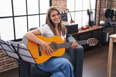 Müzik stüdyosunda klasik gitar çalan genç ve güzel bir kadın pozitif ve mutlu görünüyor ve kendine güvenen bir gülümsemeyle dişlerini gösteriyor. 