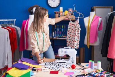 Young caucasian woman tailor holding t shirt writing on notebook at sewing studio