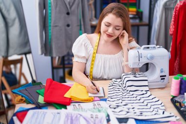 Young redhead woman tailor smiling confident writing on notebook at clothing factory