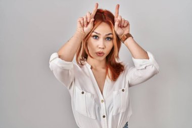 Young caucasian woman standing over isolated background doing funny gesture with finger over head as bull horns 