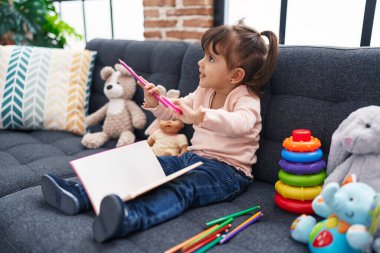 Adorable hispanic girl drawing on notebook sitting on sofa at home