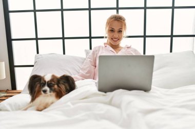 Genç beyaz kadın, yatak odasında köpekle birlikte yatağında dizüstü bilgisayar kullanıyor.
