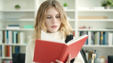 Young blonde woman student reading book standing at library university