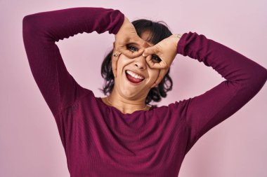 Young beautiful woman standing over pink background doing ok gesture like binoculars sticking tongue out, eyes looking through fingers. crazy expression. 