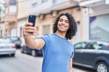 Genç Latin adam gülümsüyor. Kendine güveni tam. Sokakta akıllı telefondan selfie çekiyor.