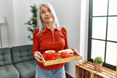 Orta yaşlı, gri saçlı, gülümseyen, elinde kahvaltı tepsisi tutan bir kadın.