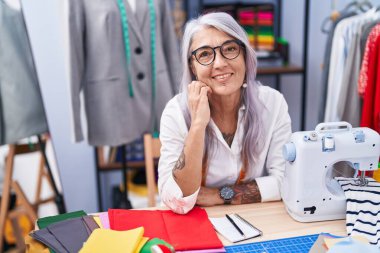 Orta yaşlı, gri saçlı, terzi dükkanında kendine güvenen, gülümseyen bir kadın.