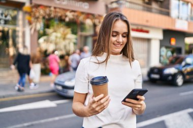 Young blonde woman using smartphone and drinking coffee at street