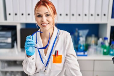 Beyaz kadın bilim adamı gülümsüyor. Laboratuvarda kendine güvenen bir gözlük tutuyor.