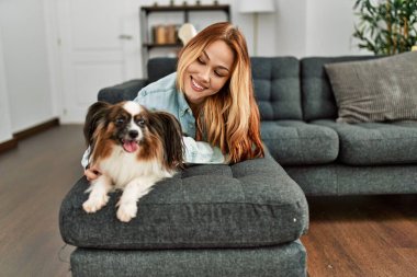 Beyaz bir kadın, evinde köpekle kanepede uzanmış kendinden emin bir şekilde gülümsüyor.