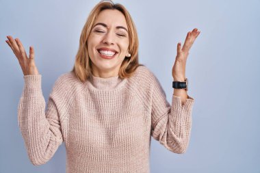 Hispanic woman standing over blue background celebrating mad and crazy for success with arms raised and closed eyes screaming excited. winner concept 