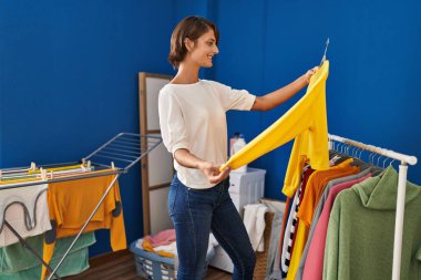 Young beautiful hispanic woman smiling confident holding sweater on clothes rack at laundry room