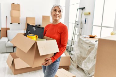 Orta yaşlı, kır saçlı, gülümseyen bir kadın yeni evinde kendine güvenen bir paket tutuyor.