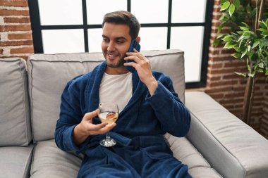Young hispanic man talking on the smartphone and drinking wine at home