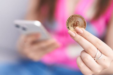 Young caucasian woman using smartphone holding uniswap coin at home clipart