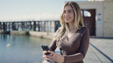 Young blonde woman smiling confident using smartphone at seaside