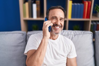 Orta yaşlı bir adam akıllı telefonda konuşuyor. Evdeki kanepede oturuyor.