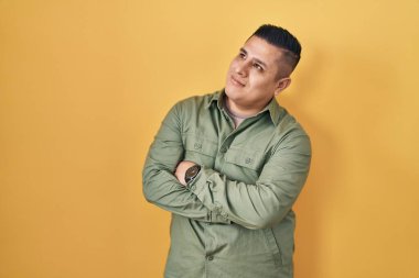 Sarı arka planda duran İspanyol genç adam kollarını kavuşturmuş ve kendinden emin bir şekilde yan tarafa bakıyor. 
