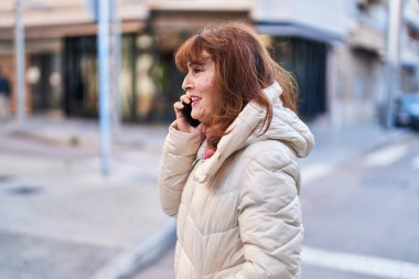 Orta yaşlı bir kadın gülümsüyor. Sokakta akıllı telefondan konuşuyor.