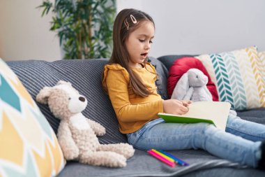 Adorable hispanic girl drawing on notebook sitting on sofa at home
