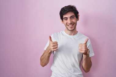 Young hispanic man standing over pink background success sign doing positive gesture with hand, thumbs up smiling and happy. cheerful expression and winner gesture. 
