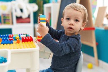 Adorable blond toddler playing with construction blocks sitting on table at kindergarten clipart