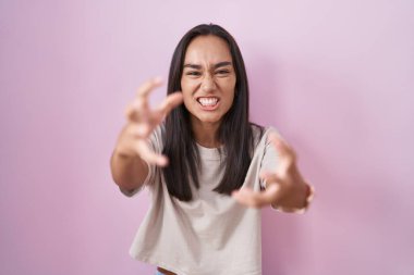 Young hispanic woman standing over pink background shouting frustrated with rage, hands trying to strangle, yelling mad 