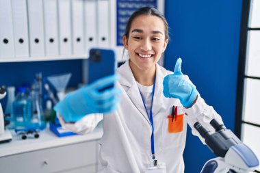 Genç Güney Asyalı kadın, akıllı telefonlu bir bilim adamının laboratuvarında çalışıyor mutlu ve pozitif gülümsüyor, başparmağıyla mükemmel ve onay işareti yapıyor. 