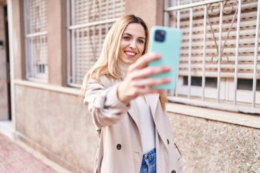 Young blonde woman smiling confident making selfie by the smartphone at street