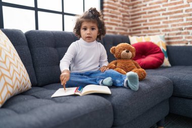 Adorable hispanic girl drawing on notebook sitting on sofa at home