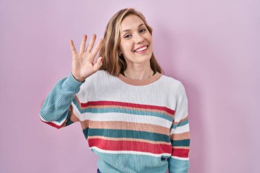 Young blonde woman standing over pink background waiving saying hello happy and smiling, friendly welcome gesture 