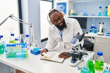 Genç Afrikalı Amerikalı adam bilim adamı üniforması giymiş kitap okuyor laboratuarda müzik dinliyor.