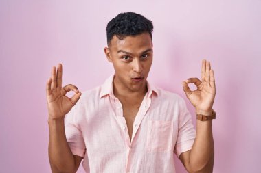 Young hispanic man standing over pink background looking surprised and shocked doing ok approval symbol with fingers. crazy expression 