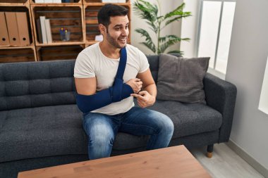 Sakallı, kol askısı takan genç İspanyol adam terapide danışmanlık yaparken mutlu bir şekilde eli ve parmağı ile işaret ediyor. 