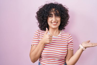 Orta Doğulu genç bir kadın pembe arka planda duruyor. Avuç içini gösteriyor ve baş parmaklarıyla iyi bir jest yapıyor. Mutlu ve neşeli gülümsüyor. 