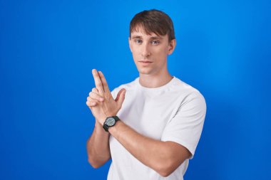 Beyaz, sarışın bir adam mavi arka planda duruyor. Elinde sembolik bir silah var. El hareketleriyle cinayet silahlarıyla oynuyor. 