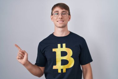 Beyaz, sarışın, bitcoin tişörtlü, neşeli, yüzünde bir gülümseme olan, mutlu ve doğal bir ifadeyle elini ve parmağını kenarı gösteren bir adam. 