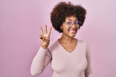 Genç Afro-Amerikalı kadın pembe arka planda duruyor ve üç numaralı parmaklarıyla işaret ederken kendinden emin ve mutlu bir şekilde gülümsüyor.. 