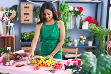 Young hispanic woman florist make bouquet of flowers at florist shop