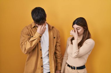 Sarı arka planda duran genç İspanyol çift yorgun burunlarını ve gözlerini ovuşturup yorgunluk ve baş ağrısı hissediyorlar. Stres ve hüsran kavramı. 