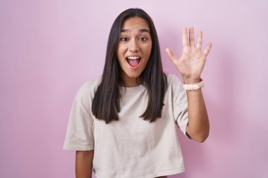 Young hispanic woman standing over pink background showing and pointing up with fingers number five while smiling confident and happy. 