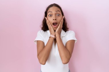 Young hispanic woman wearing casual white t shirt afraid and shocked, surprise and amazed expression with hands on face 