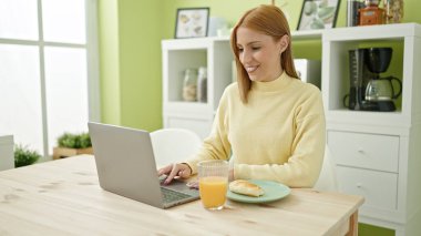Young blonde woman using laptop having breakfast at home