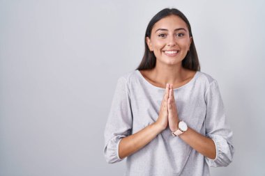 Young hispanic woman standing over white background praying with hands together asking for forgiveness smiling confident. 