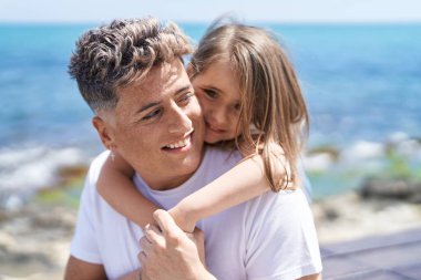 Baba ve kızı, deniz kenarında bir arada durarak gülümsüyor.